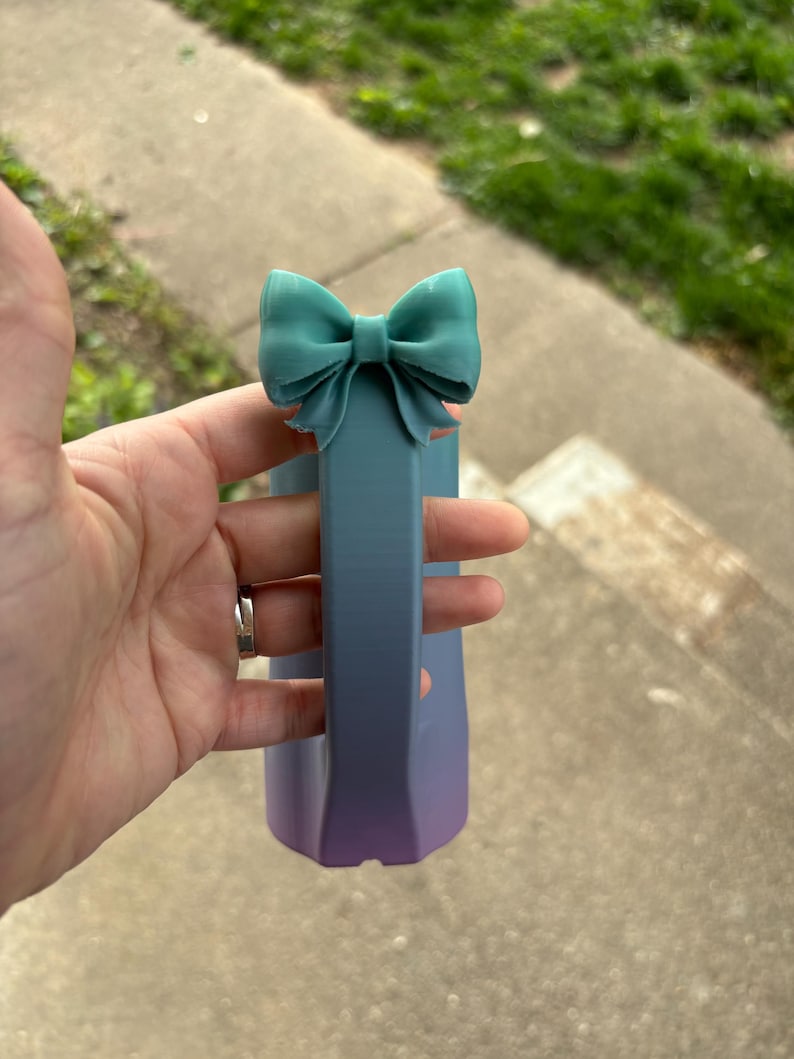 May include: A teal bow-shaped accent tops a gradient-colored, rectangular object. The object transitions from blue to purple. The bow is a solid color, while the object has a smooth, matte finish. The object's shape is geometric.