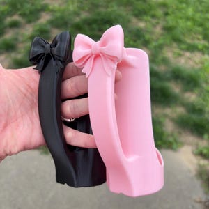 May include: Two decorative mug handles, one black and one pink, each featuring a bow at the top. The handles are held up against a blurred background of green grass and a sidewalk.