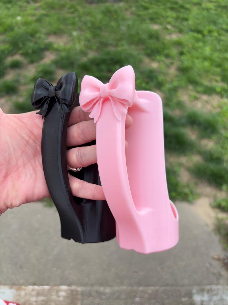 May include: Two decorative mug handles, one black and one pink, each featuring a bow at the top. The handles are held up against a blurred background of green grass and a sidewalk.