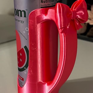 May include: A red 3D-printed can holder with a bow detail, holding a can of watermelon strawberry water. The can is red and silver with the text "om energy".