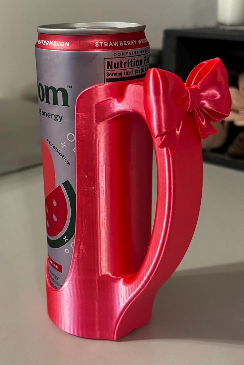 May include: A red 3D-printed can holder with a bow detail, holding a can of watermelon strawberry water. The can is red and silver with the text "om energy".