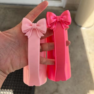 May include: Two pink bow-shaped hair accessories. One is a lighter shade of pink, and the other is a vibrant, hot pink. Both have a rectangular base. The bows are at the top of the rectangular base.