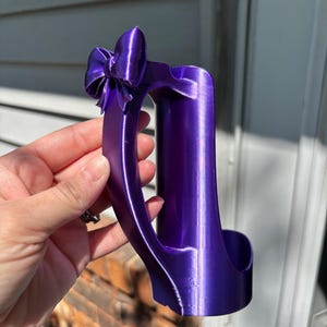 May include: A vibrant purple wine bottle holder with a bow detail. The holder is designed to cradle a wine bottle, featuring a curved handle and a decorative bow at the top. The holder is made of a shiny, smooth material.