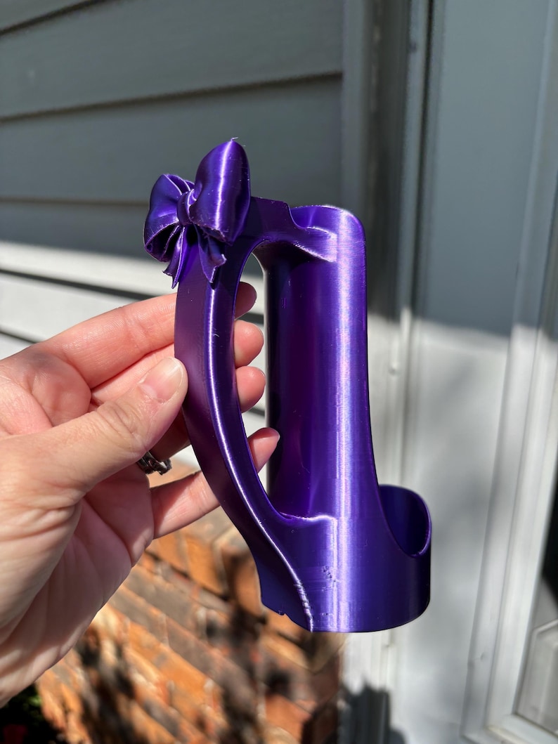 May include: A vibrant purple wine bottle holder with a bow detail. The holder is designed to cradle a wine bottle, featuring a curved handle and a decorative bow at the top. The holder is made of a shiny, smooth material.
