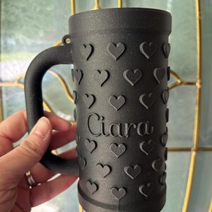 K&ouml;nnte beinhalten: Schwarzer Bierkrug mit Henkel, verziert mit gepr&auml;gten Herzen. Der Name "Ciara" ist vertikal auf der Vorderseite aufgedruckt. Der Krug wird von einer Person gehalten.