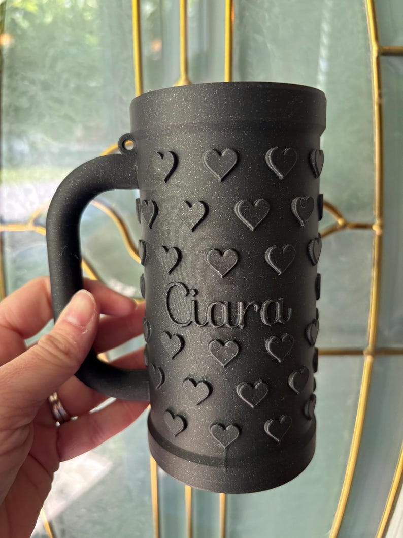 K&ouml;nnte beinhalten: Schwarzer Bierkrug mit Henkel, verziert mit gepr&auml;gten Herzen. Der Name "Ciara" ist vertikal auf der Vorderseite aufgedruckt. Der Krug wird von einer Person gehalten.