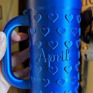K&ouml;nnte beinhalten: Eine blaue Tasse mit Henkel, verziert mit Herzformen und dem Wort "April". Die Tasse besteht aus einem einfarbigen blauen Material und hat eine zylindrische Form.