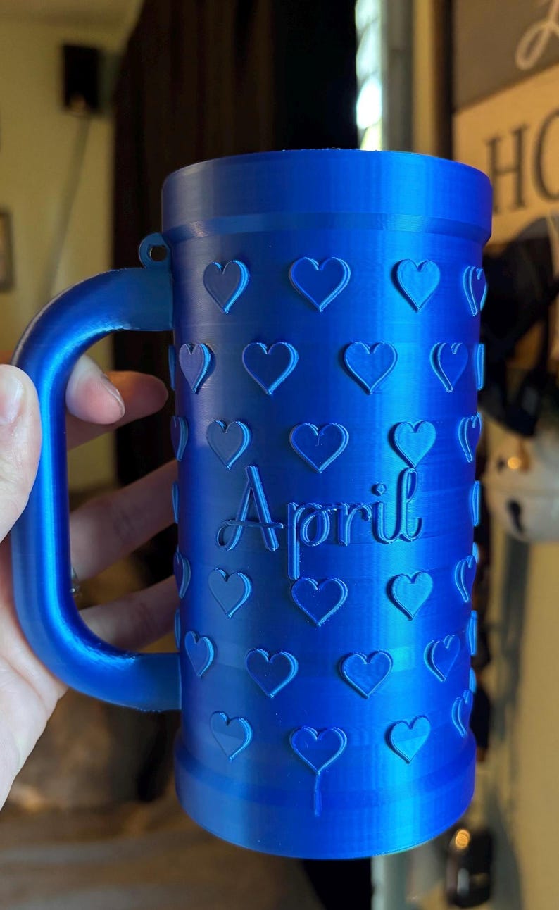 K&ouml;nnte beinhalten: Eine blaue Tasse mit Henkel, verziert mit Herzformen und dem Wort "April". Die Tasse besteht aus einem einfarbigen blauen Material und hat eine zylindrische Form.