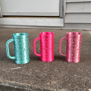 K&ouml;nnte beinhalten: Drei personalisierte Becher in T&uuml;rkis, Pink und Hellrosa, jeweils mit Henkel und herzf&ouml;rmigen Aussparungen. Auf dem t&uuml;rkisfarbenen Becher steht "Heather", auf dem pinkfarbenen Becher "Jeanie" und auf dem hellrosa Becher "Hancee".
