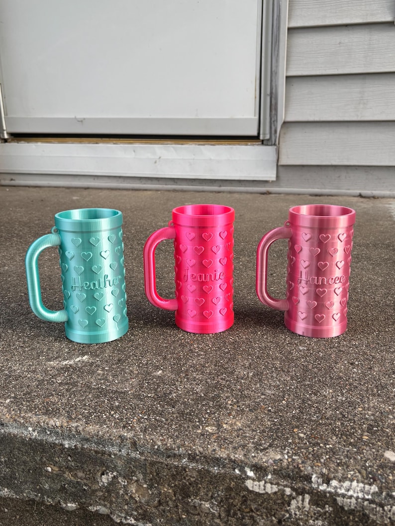 K&ouml;nnte beinhalten: Drei personalisierte Becher in T&uuml;rkis, Pink und Hellrosa, jeweils mit Henkel und herzf&ouml;rmigen Aussparungen. Auf dem t&uuml;rkisfarbenen Becher steht "Heather", auf dem pinkfarbenen Becher "Jeanie" und auf dem hellrosa Becher "Hancee".