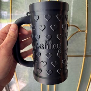 K&ouml;nnte beinhalten: Schwarzer Becher mit Henkel, verziert mit gepr&auml;gten Herzen. Der Name "Ashley" ist auf der Seite des Bechers eingraviert. Der Becher wird vor einem Fenster gehalten.