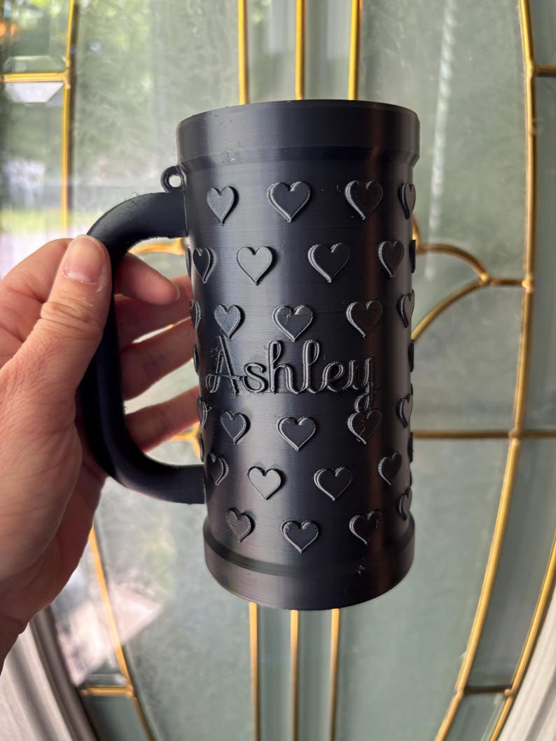 K&ouml;nnte beinhalten: Schwarzer Becher mit Henkel, verziert mit gepr&auml;gten Herzen. Der Name "Ashley" ist auf der Seite des Bechers eingraviert. Der Becher wird vor einem Fenster gehalten.