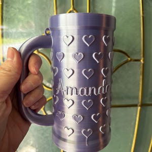 K&ouml;nnte beinhalten: Ein lavendelfarbener Becher mit Henkel, verziert mit herzf&ouml;rmigen Aussparungen. Der Name "Amanda" ist vertikal auf dem Becher aufgedruckt. Der Becher wird von einer Person gehalten.