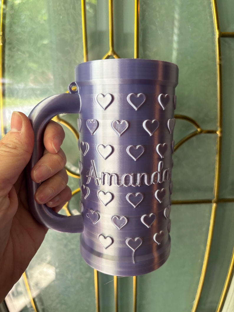 K&ouml;nnte beinhalten: Ein lavendelfarbener Becher mit Henkel, verziert mit herzf&ouml;rmigen Aussparungen. Der Name "Amanda" ist vertikal auf dem Becher aufgedruckt. Der Becher wird von einer Person gehalten.