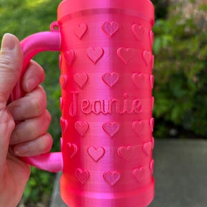 K&ouml;nnte beinhalten: Ein leuchtend rosa Becher mit Henkel, verziert mit herzf&ouml;rmigen Designs. Der Name "Jeanie" ist auf der Oberfl&auml;che des Bechers geschrieben. Der Becher ist zylindrisch und scheint aus Kunststoff zu sein.