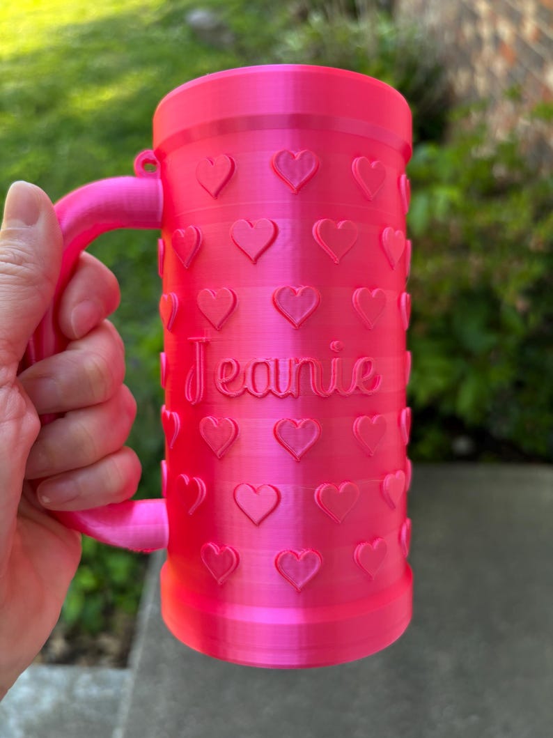 K&ouml;nnte beinhalten: Ein leuchtend rosa Becher mit Henkel, verziert mit herzf&ouml;rmigen Designs. Der Name "Jeanie" ist auf der Oberfl&auml;che des Bechers geschrieben. Der Becher ist zylindrisch und scheint aus Kunststoff zu sein.