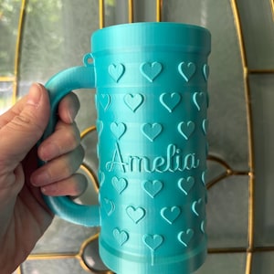 K&ouml;nnte beinhalten: Eine t&uuml;rkisfarbene Tasse mit Henkel, verziert mit gepr&auml;gten Herzen. Der Name "Amelia" ist auf der Vorderseite geschrieben. Die Tasse ist zylindrisch und scheint 3D-gedruckt zu sein.