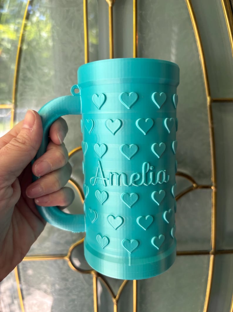 K&ouml;nnte beinhalten: Eine t&uuml;rkisfarbene Tasse mit Henkel, verziert mit gepr&auml;gten Herzen. Der Name "Amelia" ist auf der Vorderseite geschrieben. Die Tasse ist zylindrisch und scheint 3D-gedruckt zu sein.