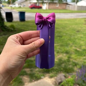 May include: A purple decorative bow with a matching purple base. The bow is a vibrant magenta color and is attached to the top of the base. The object is held in front of a blurred outdoor background.