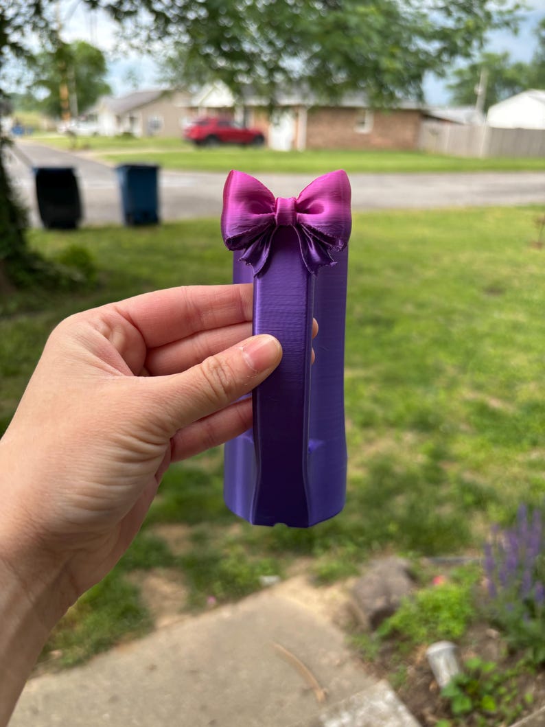 May include: A purple decorative bow with a matching purple base. The bow is a vibrant magenta color and is attached to the top of the base. The object is held in front of a blurred outdoor background.