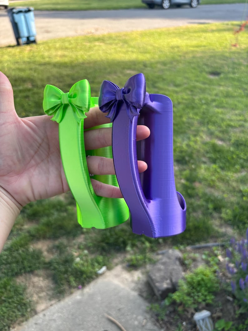 May include: Two decorative, 3D-printed handles in bright colors. One is lime green, the other is purple. Each handle features a bow at the top. The handles are designed with a curved shape and a grip.
