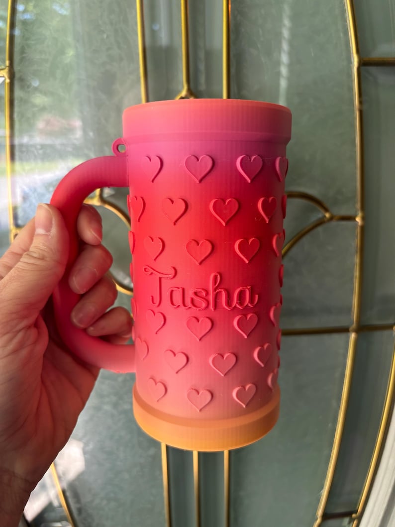 K&ouml;nnte beinhalten: Ombre-farbener Becher mit Henkel, von Rosa nach Orange verlaufend. Der Becher ist mit herzf&ouml;rmigen Aussparungen und dem Namen "Tasha" verziert. Personalisierte Trinkbecher.