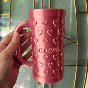 K&ouml;nnte beinhalten: Eine rosa Tasse mit Henkel, verziert mit herzf&ouml;rmigen Aussparungen. Der Name "Hancee" ist in Schreibschrift auf die Tasse gedruckt. Die Tasse besteht aus einem festen Material und hat eine zylindrische Form.