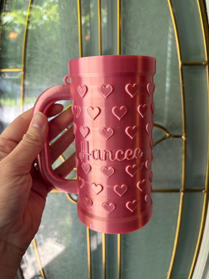 K&ouml;nnte beinhalten: Eine rosa Tasse mit Henkel, verziert mit herzf&ouml;rmigen Aussparungen. Der Name "Hancee" ist in Schreibschrift auf die Tasse gedruckt. Die Tasse besteht aus einem festen Material und hat eine zylindrische Form.