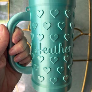 K&ouml;nnte beinhalten: Eine t&uuml;rkisfarbene Tasse mit Henkel, verziert mit herzf&ouml;rmigen Designs. Der Name "Heather" ist auf der Oberfl&auml;che der Tasse eingraviert. Die zylindrische Tasse scheint aus einem 3D-gedruckten Material zu bestehen.