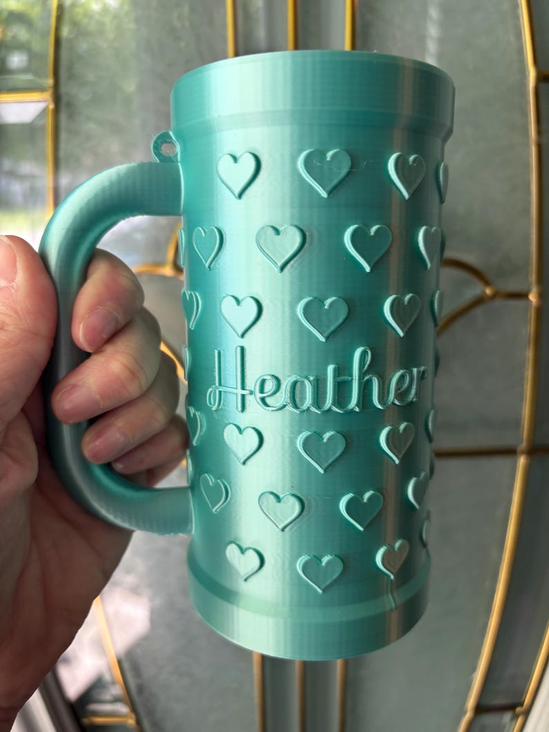 K&ouml;nnte beinhalten: Eine t&uuml;rkisfarbene Tasse mit Henkel, verziert mit herzf&ouml;rmigen Designs. Der Name "Heather" ist auf der Oberfl&auml;che der Tasse eingraviert. Die zylindrische Tasse scheint aus einem 3D-gedruckten Material zu bestehen.