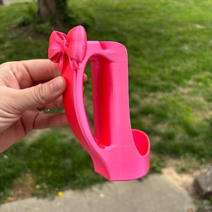 May include: A bright pink, 3D-printed phone holder with a bow detail. The holder features a curved handle and a slot to hold a phone. The design is a solid, vibrant pink color.