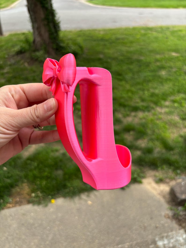 May include: A bright pink, 3D-printed phone holder with a bow detail. The holder features a curved handle and a slot to hold a phone. The design is a solid, vibrant pink color.