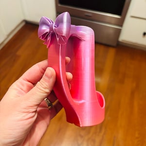 May include: A pink and purple high heel-shaped phone holder with a bow on top. The holder is made of a 3D-printed material and has a gradient color scheme, transitioning from pink to purple.