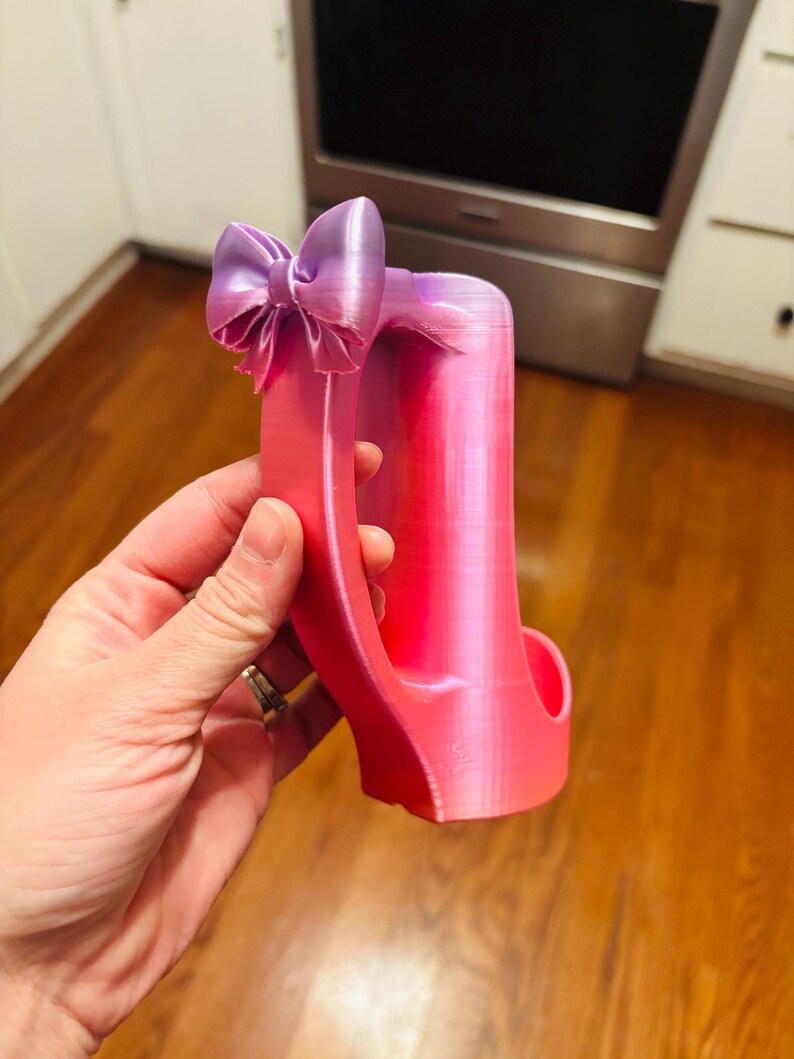 May include: A pink and purple high heel-shaped phone holder with a bow on top. The holder is made of a 3D-printed material and has a gradient color scheme, transitioning from pink to purple.