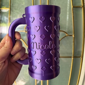 K&ouml;nnte beinhalten: Ein lila Becher mit Henkel, verziert mit Herzformen. Der Name "Nicole" ist vertikal auf dem Becher aufgedruckt. Der Becher wird in der Hand einer Person gehalten.