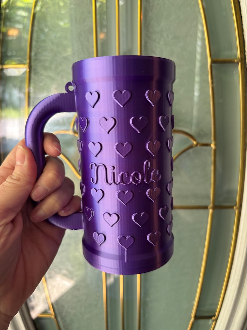 K&ouml;nnte beinhalten: Ein lila Becher mit Henkel, verziert mit Herzformen. Der Name "Nicole" ist vertikal auf dem Becher aufgedruckt. Der Becher wird in der Hand einer Person gehalten.