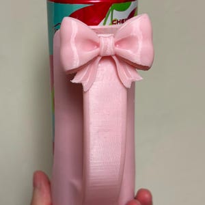 May include: A pink, 3D-printed can cooler with a bow handle. The handle is a rectangular shape with a bow at the top. The can cooler is designed to hold a standard-sized beverage can. The can inside has a red and teal label.
