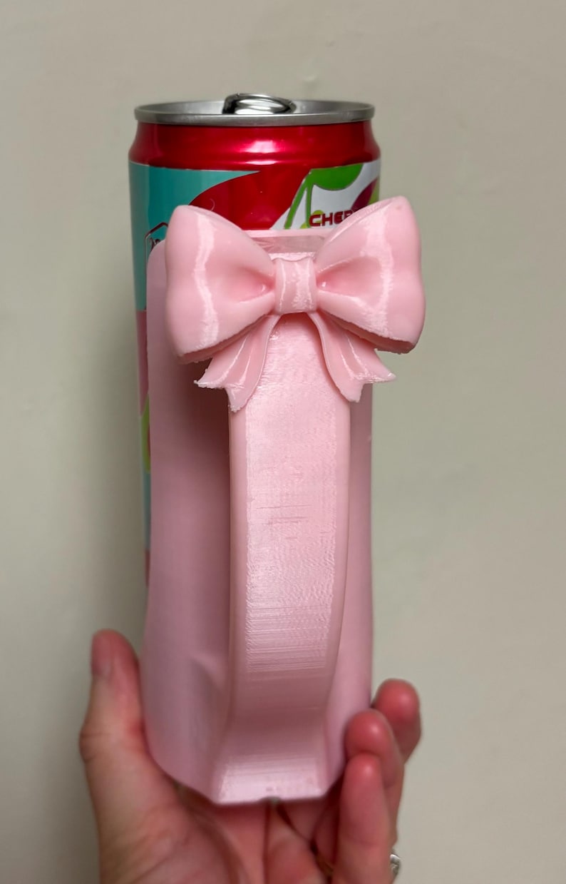May include: A pink, 3D-printed can cooler with a bow handle. The handle is a rectangular shape with a bow at the top. The can cooler is designed to hold a standard-sized beverage can. The can inside has a red and teal label.