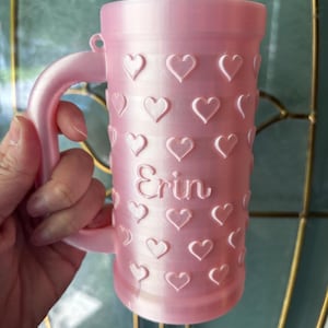 K&ouml;nnte beinhalten: Eine rosa Tasse mit Henkel, verziert mit gepr&auml;gten Herzen. Der Name "Erin" ist auf die Tasse gedruckt. Die Tasse besteht aus einem festen Material und hat eine glatte Oberfl&auml;che. Die Tasse ist f&uuml;r Getr&auml;nke konzipiert.