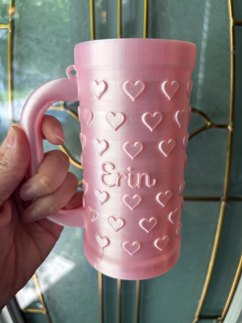 K&ouml;nnte beinhalten: Eine rosa Tasse mit Henkel, verziert mit gepr&auml;gten Herzen. Der Name "Erin" ist auf die Tasse gedruckt. Die Tasse besteht aus einem festen Material und hat eine glatte Oberfl&auml;che. Die Tasse ist f&uuml;r Getr&auml;nke konzipiert.