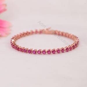 Könnte beinhalten: Ein Tennisarmband aus Roségold mit einer durchgehenden Reihe herzförmiger, leuchtend rosaroter Edelsteine. Die Metallfassung ist poliert und glänzend, und der Schriftzug Artist Jewels Boutique ist in der Mitte des Armbands zu sehen.