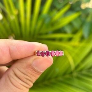 Könnte beinhalten: Ein goldfarbener Ring mit einer Reihe von oval geschliffenen, tiefroten Edelsteinen. Die Edelsteine sind eng aneinander gesetzt und bilden ein durchgehendes Farbband. Der Ring wird vor einem Hintergrund aus grünem Laubwerk präsentiert, das das leuchtende Rot der Steine hervorhebt.