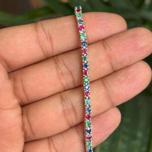 May include: A silver bracelet with a row of alternating colorful gemstones, including red, green, and blue stones. The bracelet is held against a blurred green background, showcasing the vibrant colors of the gems.