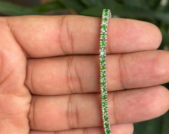 Puede incluir: Una delicada pulsera de plata con una fila de gemas verdes vibrantes. Las gemas están espaciadas uniformemente y engastadas en una banda de plata. La pulsera se muestra sobre un fondo verde borroso.