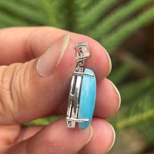 May include: A silver pendant featuring a rectangular, light blue gemstone. The pendant has a square bail and a decorative silver frame. The background is a blurred green.