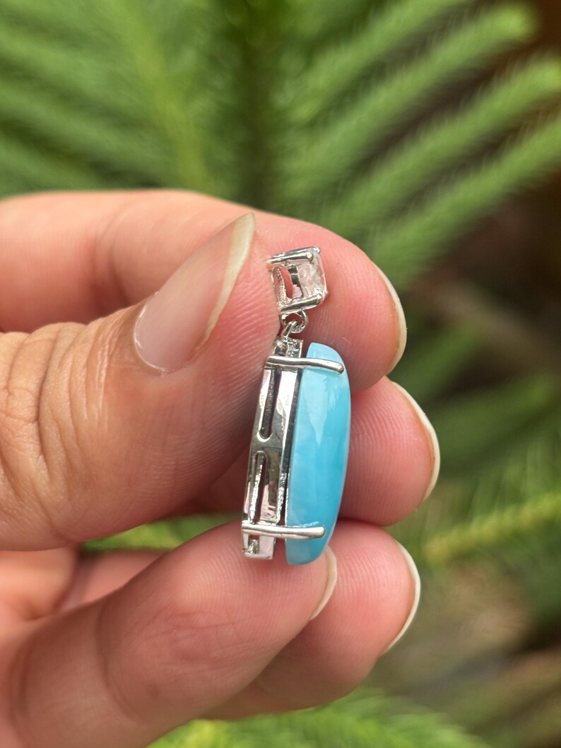 May include: A silver pendant featuring a rectangular, light blue gemstone. The pendant has a square bail and a decorative silver frame. The background is a blurred green.