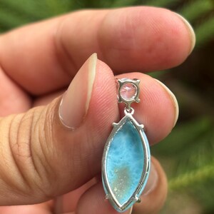 May include: A silver pendant featuring a light blue, oval gemstone and a smaller, pink, round stone. The pendant is held by a hand against a blurred green backdrop. The blue stone has a distinctive pattern.