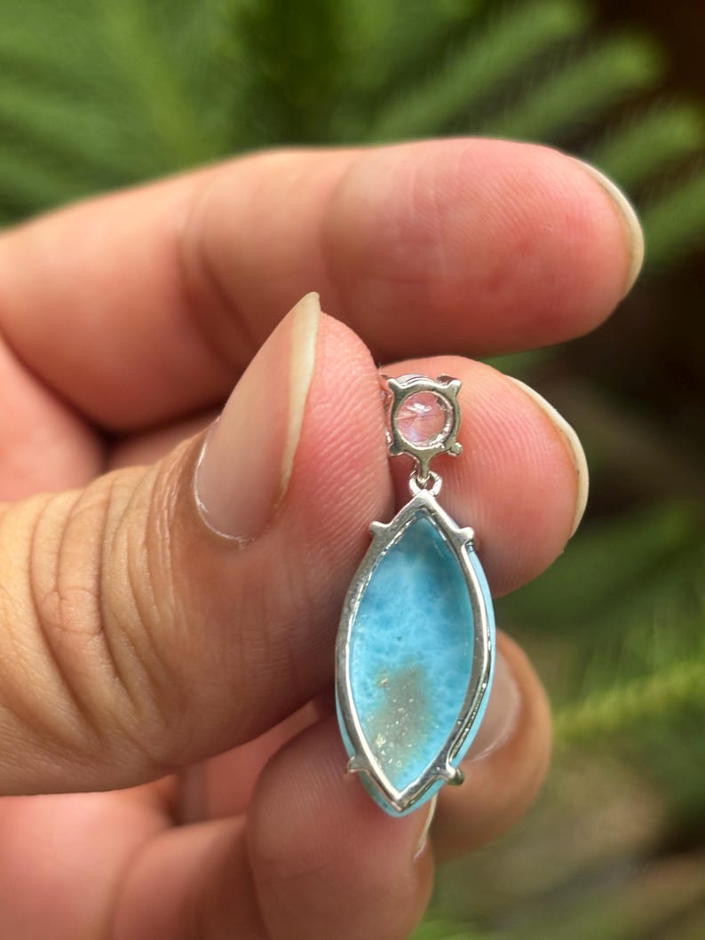 May include: A silver pendant featuring a light blue, oval gemstone and a smaller, pink, round stone. The pendant is held by a hand against a blurred green backdrop. The blue stone has a distinctive pattern.