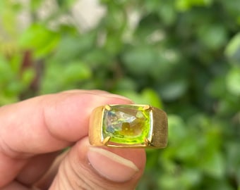 May include: A gold ring featuring a large, rectangular, light green gemstone. The ring has a brushed gold band and a bezel setting that holds the gemstone securely. The background is a blurred view of green foliage.