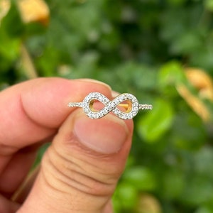 Könnte beinhalten: Ein zarter Ring mit einem Unendlichkeitszeichen. Das Unendlichkeitszeichen ist mit kleinen, funkelnden Steinen besetzt, die in einem goldfarbenen Band gefasst sind. Der Ring wird vor einem verschwommenen grünen Hintergrund gehalten.